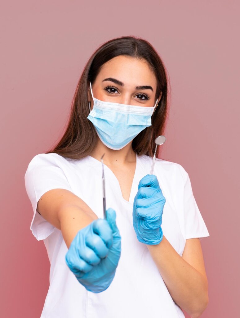 A dental assistant wearing a mask and gloves holds dental tools in front of a pink background.