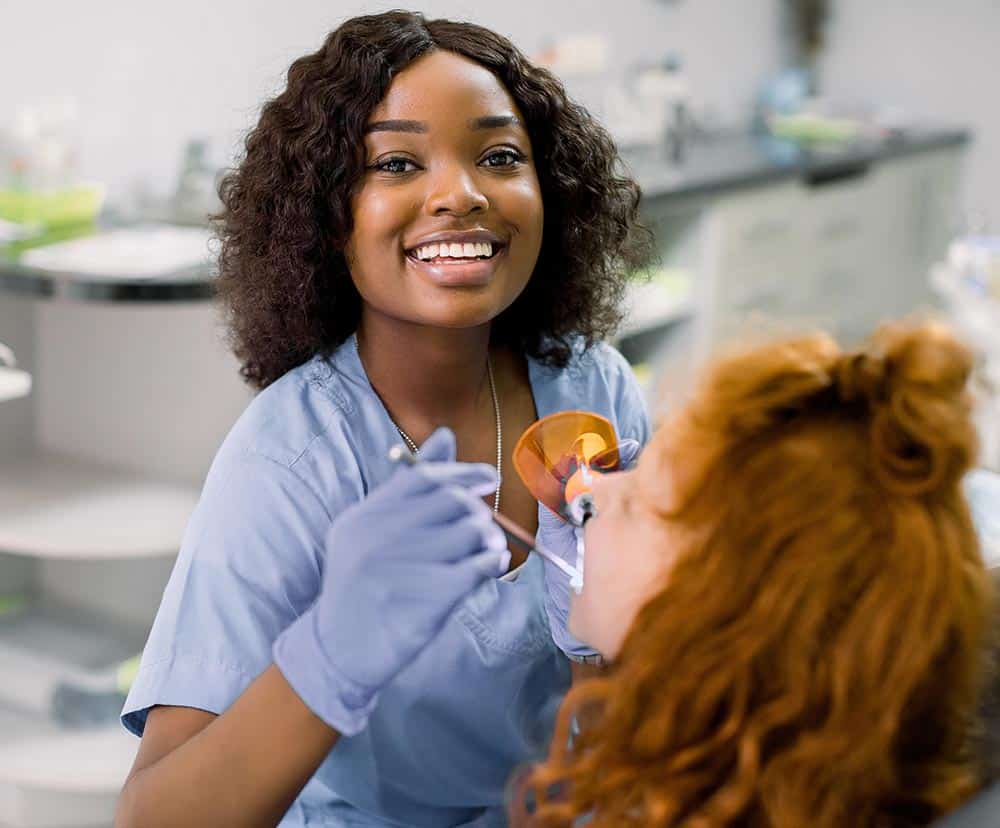 A dentist wearing blue scrubs and gloves smiles while treating a patient with reddish hair, assisted by a diligent dental assistant, in a bright, welcoming dental clinic.